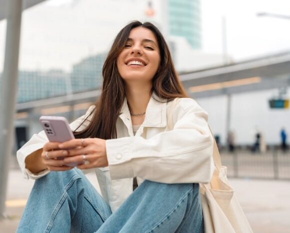 Frau sitzt am Bahnhof und schaut lächelnd vom Handy hoch