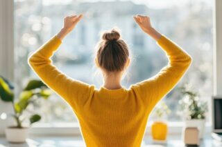 Eine Person in einem gelben Pullover streckt sich mit erhobenen Armen vor einem Fenster. Sonnenlicht erhellt den Raum, auf dessen Fensterbank grüne Pflanzen und ein kleiner Topf stehen. Dies schafft eine Atmosphäre, die dazu einlädt, den friedlichen Morgen zu genießen.
