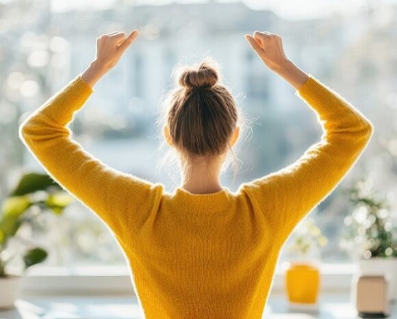 Eine Person in einem gelben Pullover streckt sich mit erhobenen Armen vor einem Fenster. Sonnenlicht erhellt den Raum, auf dessen Fensterbank grüne Pflanzen und ein kleiner Topf stehen. Dies schafft eine Atmosphäre, die dazu einlädt, den friedlichen Morgen zu genießen.