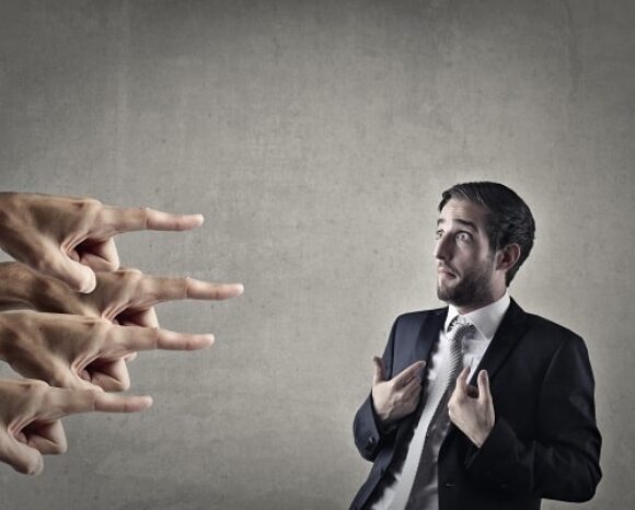 Ein Mann im Anzug blickt überrascht und zeigt auf sich, während mehrere Hände von links vor einem grauen Hintergrund anklagend in seine Richtung weisen und so die Spannung der Schuldzuweisung verkörpern.