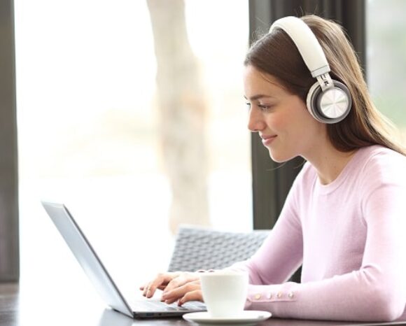 Eine Person mit Kopfhörern sitzt an einem Tisch und tippt auf einem Laptop. Eine weiße Kaffeetasse steht auf dem Tisch. Im Hintergrund ist ein Fenster mit unscharfem Blick nach draußen zu sehen.