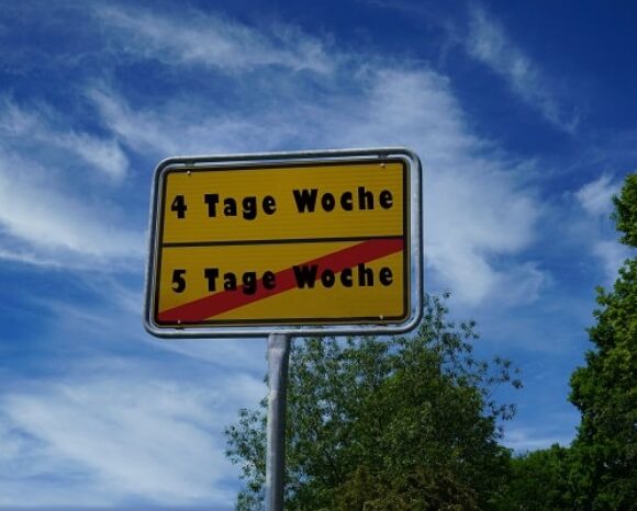 Ein gelbes Straßenschild unter blauem Himmel und weißen Wolken trägt die Aufschrift „Vier-Tage-Woche“ über der durchgestrichenen „5 Tage Woche“, die den Wechsel zur Vier-Tage-Woche symbolisiert. Am unteren Bildrand sind üppige Bäume zu sehen.