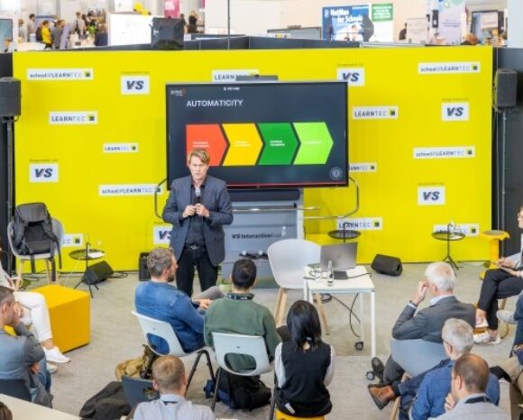 Ein Referent präsentiert an einem Konferenzstand eine Folie mit dem Titel „Automatisierung“, die auf einem Bildschirm angezeigt wird. Der Bildschirm zeigt ein Flussdiagramm mit farbigen Segmenten. Die Teilnehmer sitzen im Halbkreis. Der Bereich ist mit den Logos „VS“ und „LEARNTEC“ gekennzeichnet.