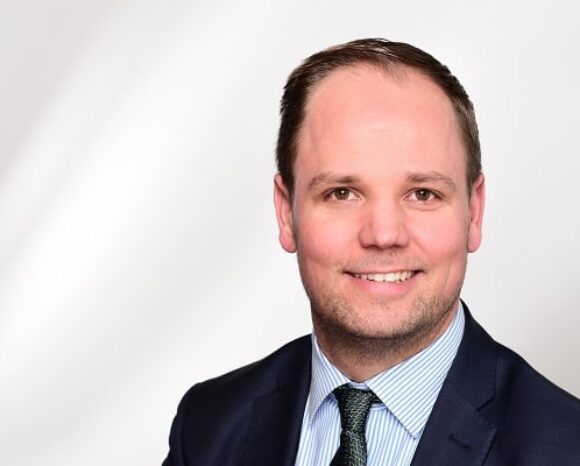 Ein Mann im dunklen Anzug mit hellblau gestreiftem Hemd und gemusterter Krawatte lächelt in die Kamera und verkörpert Professionalität in der Personalarbeit. Er hat kurze Haare und einen leichten Bart. Der Hintergrund ist schlicht und sanft beleuchtet, was eine einladende Atmosphäre schafft.