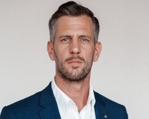 Ein Mann mit kurzen Haaren und Bart trägt ein blaues Jackett über einem weißen Hemd. Er steht vor einem schlichten weißen Hintergrund und blickt mit neutralem Gesichtsausdruck direkt in die Kamera.