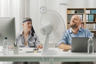 Zwei Männer schwitzen im Büro und haben den Ventilator angeschaltet. HR kann hier als Hitzebeauftragter wirken.