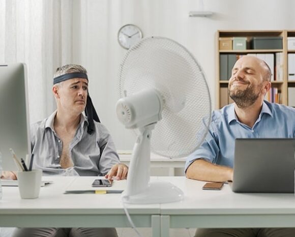 Zwei Männer schwitzen im Büro und haben den Ventilator angeschaltet. HR kann hier als Hitzebeauftragter wirken.