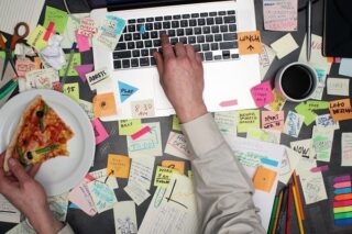 Mann arbeitet am Laptop, der voll mit Post-its ist, und isst nebenbei Pizza. Er ist scheinbar ein Workaholic.