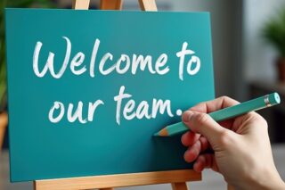 Eine Hand schreibt mit einem Stift in weiß "Welcome to our team" auf eine türkisfarbene Tafel.