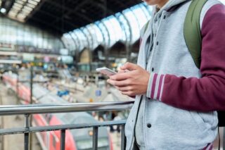 Mann steht am Bahnhof und schaut auf sein Handy mit dem Deutschlandticket.