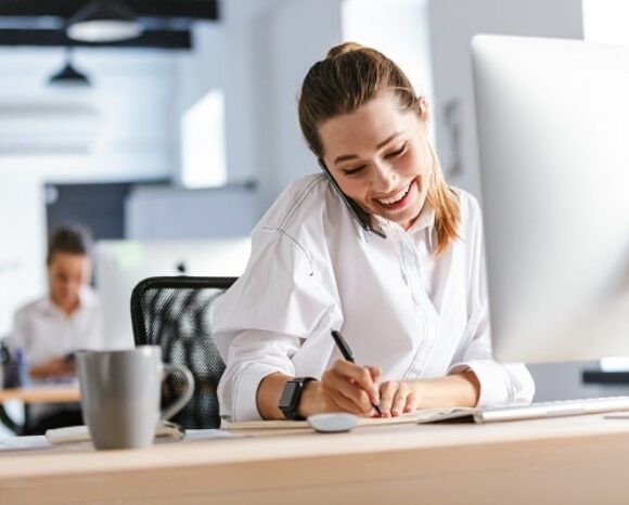 Frau telefoniert und macht Notizen am Schreibtisch im Büro
