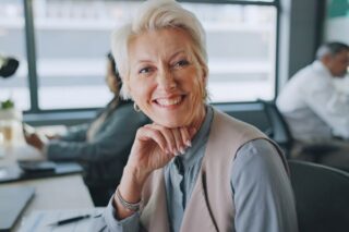 Ältere Mitarbeiterin lächelt am Arbeitsplatz in die Kamera