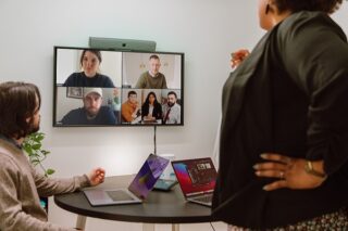 Kollegen sitzen im Büro und besprechen sich mit anderen Kollegen digital, die auf dem Bildschirm zu sehen sind.