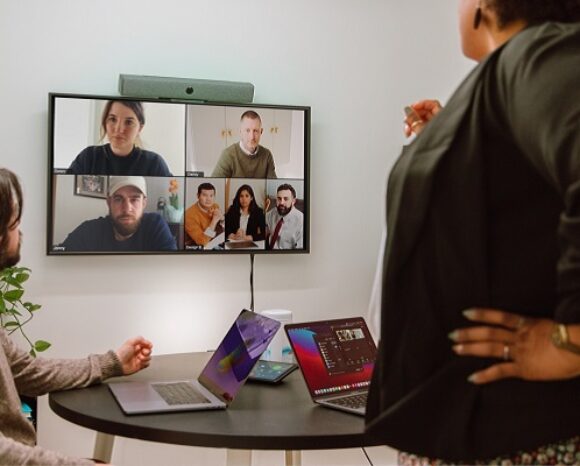 Kollegen sitzen im Büro und besprechen sich mit anderen Kollegen digital, die auf dem Bildschirm zu sehen sind.