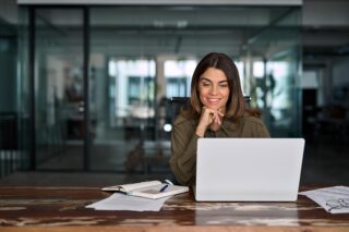 Frau sitzt lächelnd am Schreibtisch vor einem Laptop in einem modernen Büro. Neben ihr liegen Notizbuch und Unterlagen. Das Bild symbolisiert effiziente, digitale HR-Arbeit und moderne Personalprozesse.