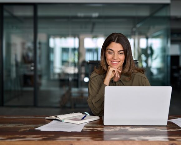 Frau sitzt lächelnd am Schreibtisch vor einem Laptop in einem modernen Büro. Neben ihr liegen Notizbuch und Unterlagen. Das Bild symbolisiert effiziente, digitale HR-Arbeit und moderne Personalprozesse.