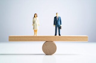 Zwei Figuren, eine Frau und ein Mann, halten sich auf einem Holzbalken die Waage.