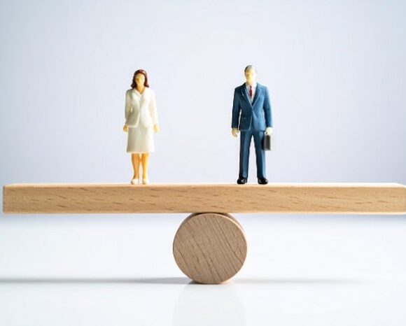Zwei Figuren, eine Frau und ein Mann, halten sich auf einem Holzbalken die Waage.