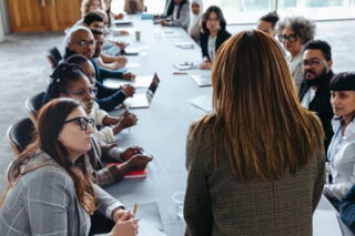 Frau steht vor Kollegen an einem langen Tisch und spricht über Personalführung
