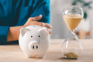 Mann sitzt am Tisch, vor ihm stehen ein Sparschwein und eine Sanduhr