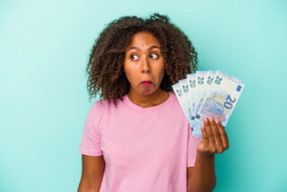 Frau mit lockigen Haaren hält Geldscheine in der Hand und macht ein unwissendes Gesicht