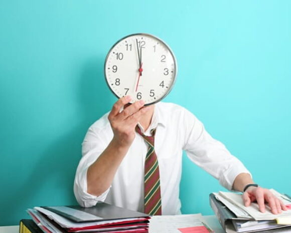 Mehrarbeit: Mann hält Uhr vors Gesicht am Schreibtisch