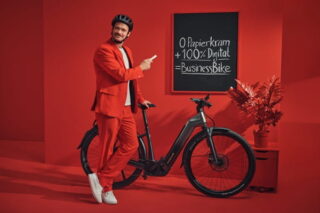 Mann in rotem Anzug steht vor roter Wand mit Fahrrad, an der eine Tafel hängt