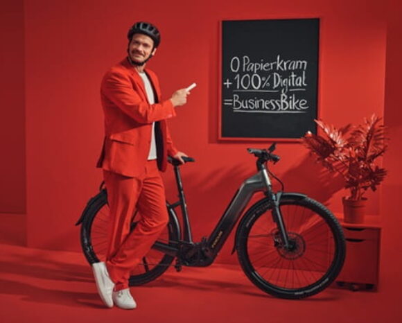 Mann in rotem Anzug steht vor roter Wand mit Fahrrad, an der eine Tafel hängt