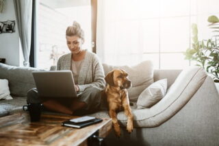 Frau mit Hund neben sich arbeitet auf dem Sofa im Homeoffice am Laptop